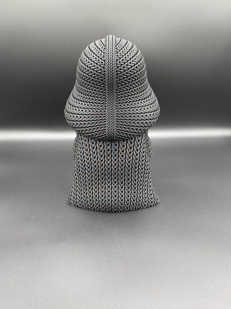 May include: A gray, textured sculpture of a figure with a rounded head and a flowing, skirt-like base. The sculpture has a woven or chainmail-like appearance, with intricate patterns across its surface. The figure is set against a dark gray background.