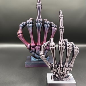 May include: Two skeleton hand sculptures, each displaying the middle finger. One is a gradient of blue, purple, and pink, while the other is silver. Both are on square bases, perfect for a unique home decor statement.