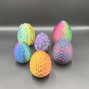 May include: A collection of seven decorative, colorful dragon eggs. Each egg has a unique design and color scheme, including blue, purple, yellow, orange, pink, and green. The eggs are arranged on a dark surface.