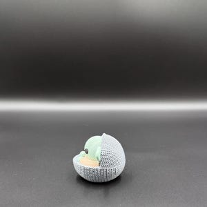 May include: A 3D-printed figurine of a character from a popular science fiction series. The figure is in a gray, knitted-style cradle, with a light green head and tan body. The background is a gradient of dark gray.