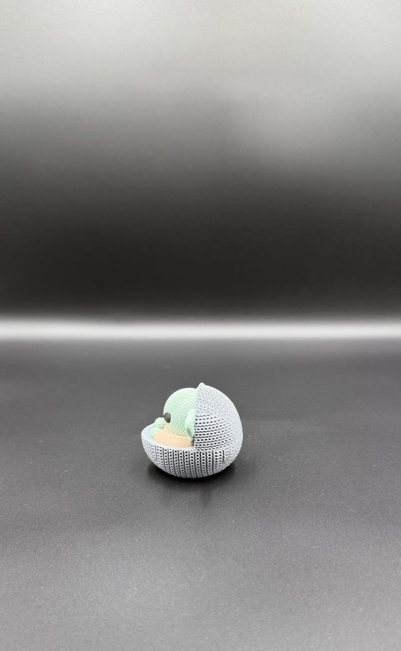 May include: A 3D-printed figurine of a character from a popular science fiction series. The figure is in a gray, knitted-style cradle, with a light green head and tan body. The background is a gradient of dark gray.