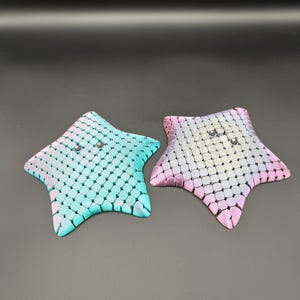 May include: Two star-shaped decorative trays. One is teal and the other is pink, both with a textured, scale-like pattern. Each has three small, silver-colored embellishments. The trays are on a dark background.
