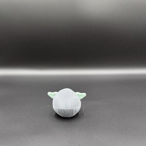 May include: A small, round, light gray knitted figure with two light green ears. The figure is set against a dark gray background. The object appears to be a toy or decorative item.