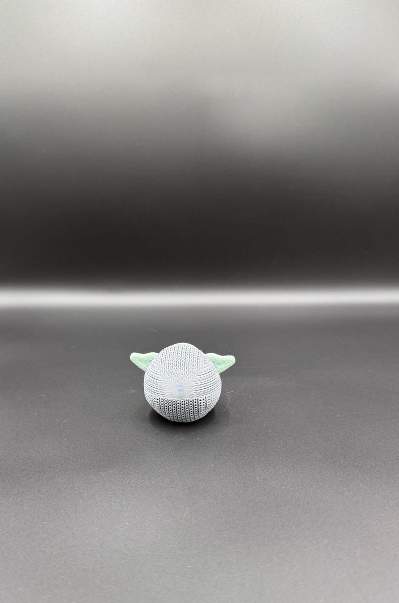 May include: A small, round, light gray knitted figure with two light green ears. The figure is set against a dark gray background. The object appears to be a toy or decorative item.