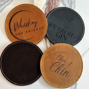 May include: Four leather coasters with different sayings. The coasters are brown and black with white text. The sayings are "Whiskey and Friends", "We Trust Gin", "Beer Dear Try Beer", and "Good Life Chin! Chin!"