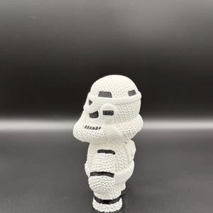 May include: A white knitted figurine of a Stormtrooper helmet. The helmet has black accents and is displayed against a dark background. The knitted texture is visible, adding detail to the design.