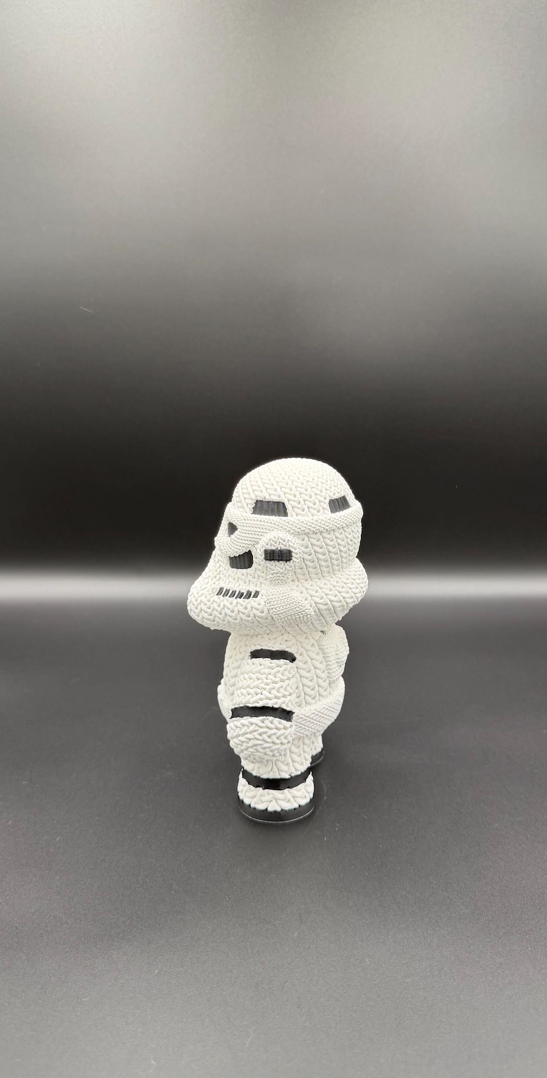 May include: A white knitted figurine of a Stormtrooper helmet. The helmet has black accents and is displayed against a dark background. The knitted texture is visible, adding detail to the design.