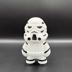 May include: A white, knitted Stormtrooper figurine with black accents. The figure has a detailed helmet design, black stripes on the arms and legs, and a textured appearance. The figurine is standing on a dark surface.