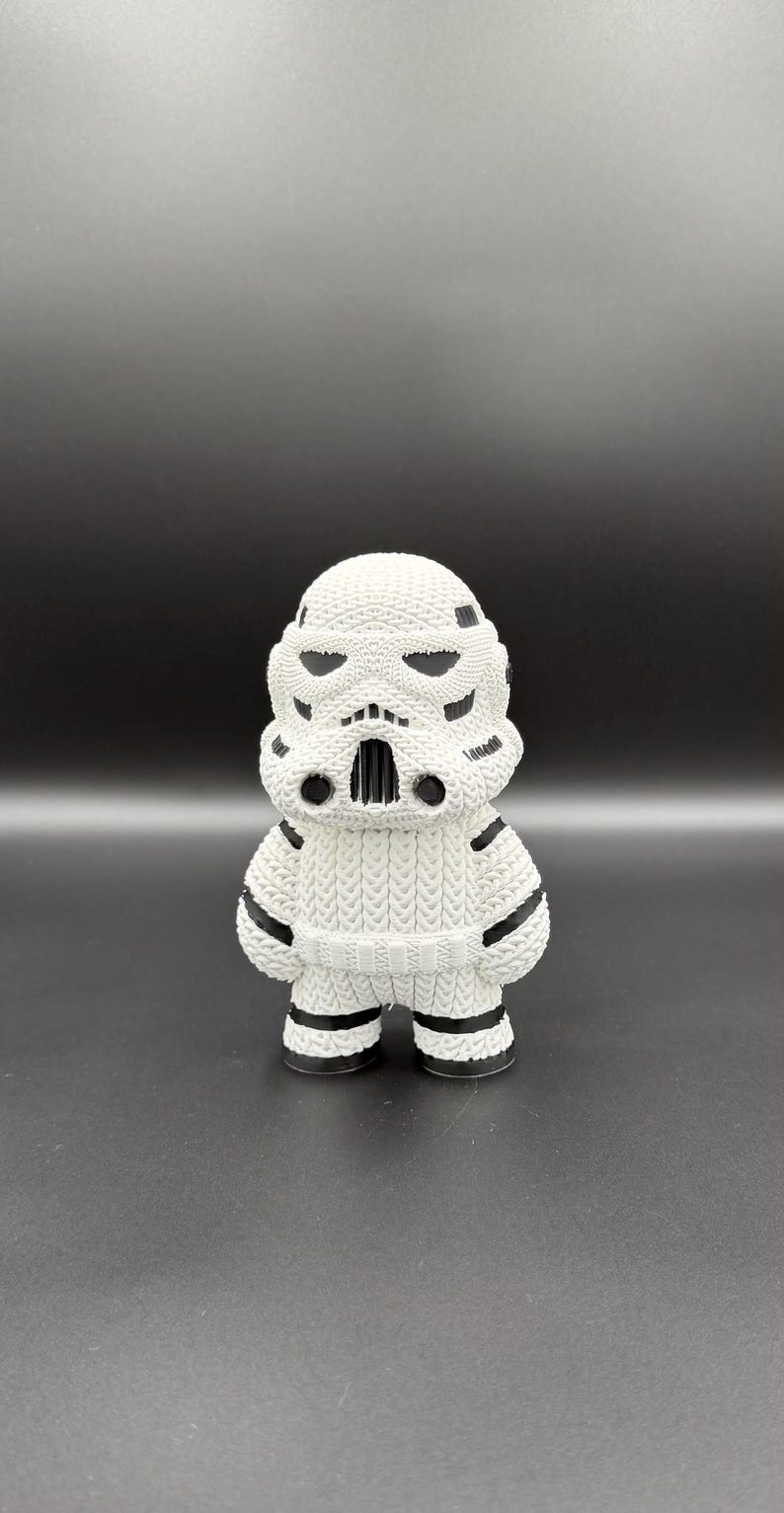 May include: A white, knitted Stormtrooper figurine with black accents. The figure has a detailed helmet design, black stripes on the arms and legs, and a textured appearance. The figurine is standing on a dark surface.