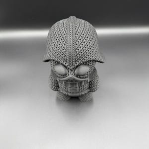 May include: A gray, textured figurine with a helmet-like head and a detailed face. The figure has a unique design, with intricate patterns covering its head and body. The overall aesthetic is reminiscent of a stylized character or sculpture.