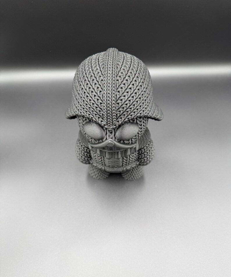 May include: A gray, textured figurine with a helmet-like head and a detailed face. The figure has a unique design, with intricate patterns covering its head and body. The overall aesthetic is reminiscent of a stylized character or sculpture.