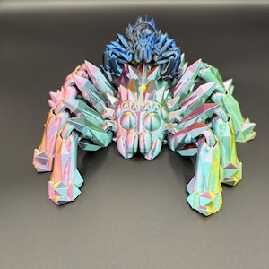 May include: A multi-colored, geometric spider sculpture with iridescent hues of pink, green, and yellow. The spider has a detailed, crystal-like design and a smaller blue spider on its back. The sculpture is set against a dark background.