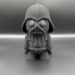 May include: A gray 3D-printed figurine of Darth Vader, featuring a knitted texture design. The figure has a helmet, mask, and chest plate, all rendered in a detailed, textured pattern. The overall aesthetic is a blend of pop culture and craft.