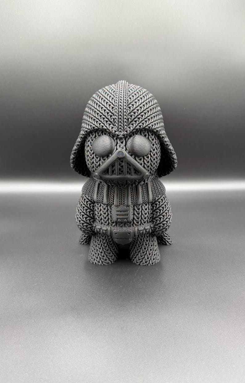 May include: A gray 3D-printed figurine of Darth Vader, featuring a knitted texture design. The figure has a helmet, mask, and chest plate, all rendered in a detailed, textured pattern. The overall aesthetic is a blend of pop culture and craft.