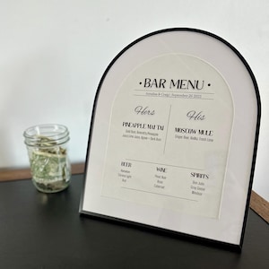 May include: A bar menu with a black frame, featuring a white background and black text. The menu lists cocktails, beer, wine, and spirits. A glass jar with green contents sits to the left of the menu.