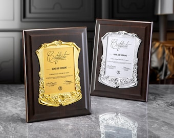 Puede incluir: Dos placas de certificado enmarcadas en madera con diseños en relieve dorado y plateado. Las placas presentan la palabra "Certificado" en una fuente de letra decorativa. La placa dorada tiene un diseño floral y la placa plateada tiene un diseño de vid.