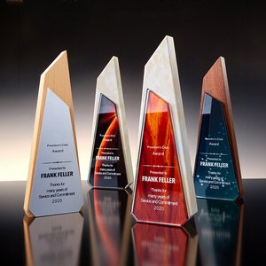 May include: Four wooden award plaques with different colored glass inserts. Each plaque has a silver metal plate with the text "President's Club Award" and "Presented to FRANK FELLER" with a message of thanks for service and commitment. The plaques are on a black reflective surface.