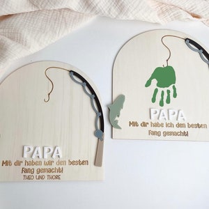 Könnte beinhalten: Zwei Holzschilder mit einem Fischthema. Die Schilder sind bogenförmig und haben einen Angelruten- und Fisch-Ausschnitt. Der Text auf den Schildern lautet "PAPA Mit dir haben wir den besten Fang gemacht!" und "PAPA Mit dir habe ich den besten Fang gemacht!"