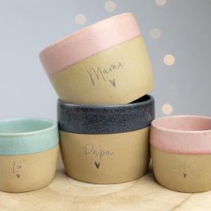 Op de afbeelding: Een stapel keramische bekers in verschillende kleuren, waaronder roze, blauw en grijs. De bekers hebben een tweekleurig ontwerp met een lichtbruine basis en een gekleurde rand. Sommige bekers hebben tekst zoals "Mama" en "Papa".