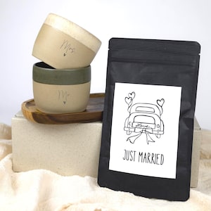 Könnte beinhalten: Zwei Keramikbecher, einer beige und einer grün, mit den Aufschriften "Mrs." und "Mr.". Eine schwarze Tüte mit einem "Just Married"-Etikett mit einer Autoillustration befindet sich neben den Bechern. Die Becher stehen auf einem Holztablett.