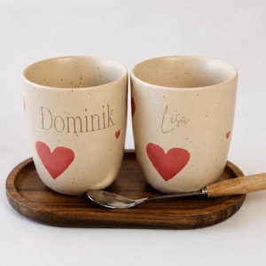 Könnte beinhalten: Zwei beige Keramikbecher mit roten Herzmotiven und den Namen "Dominik" und "Lisa" stehen auf einem Holztablett. Ein kleiner Löffel mit Holzgriff liegt auf dem Tablett. Die Becher haben eine gesprenkelte Textur.