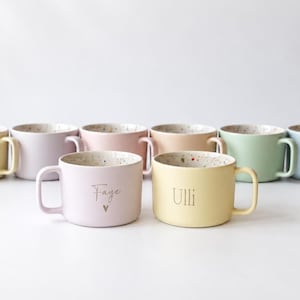 Könnte beinhalten: Eine Sammlung von acht Keramiktassen in Pastellfarben wie Gelb, Rosa, Pfirsich, Grün und Blau. Jede Tasse hat ein gesprenkeltes Inneres und eine glatte, matte Außenseite. Zwei Tassen im Vordergrund sind mit den Namen Faye und Ulli personalisiert.