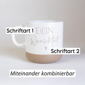 Op de afbeelding: Witte keramische mok met een gespikkelde afwerking en een bruine basis. De mok heeft de tekst "DEIN Wunschtext" in een scriptlettertype, samen met een klein hartje. De teksten "Schriftart 1" en "Schriftart 2" zijn ook zichtbaar. De tekst "Miteinander kombinierbar" staat onderaan.