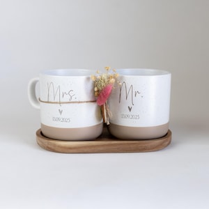 Op de afbeelding: Twee witte keramische mokken met een gespikkeld ontwerp en beige bodems staan op een houten dienblad. Op &eacute;&eacute;n mok staat "Mrs." en op de andere "Mr." met een datum eronder. Een klein boeketje droogbloemen is ertussen gebonden.