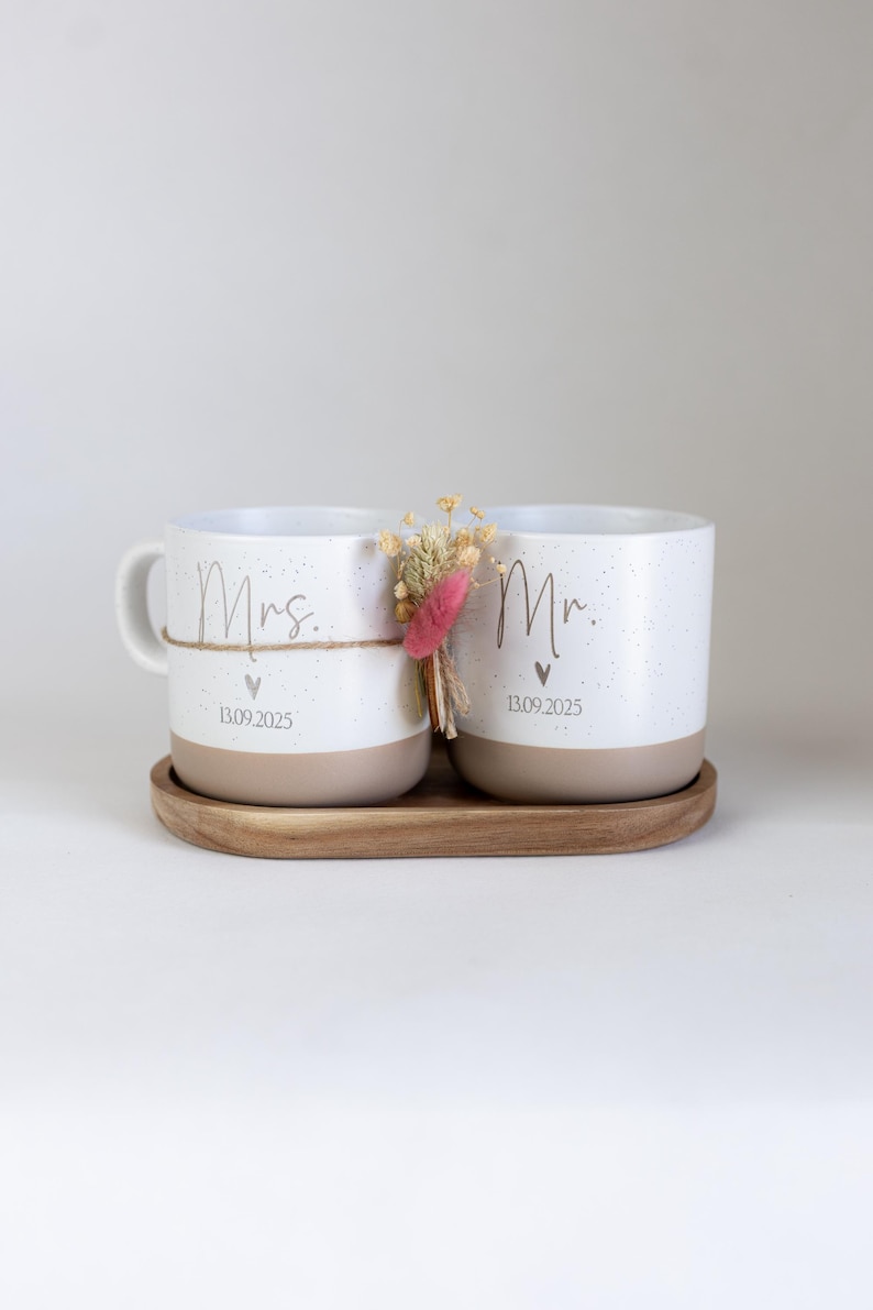 Op de afbeelding: Twee witte keramische mokken met een gespikkeld ontwerp en beige bodems staan op een houten dienblad. Op &eacute;&eacute;n mok staat "Mrs." en op de andere "Mr." met een datum eronder. Een klein boeketje droogbloemen is ertussen gebonden.