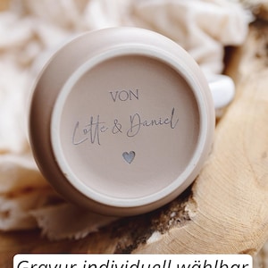 Op de afbeelding: Een beige keramische mok met de tekst "Von Lotte & Daniel" en een hartsymbool. De mok staat op een houten oppervlak. De tekst "Beispiel" en "Gravur individuell w&auml;hlbar" zijn ook zichtbaar.