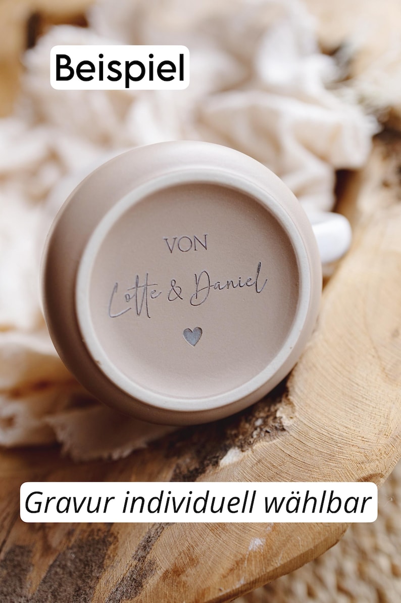 Op de afbeelding: Een beige keramische mok met de tekst "Von Lotte & Daniel" en een hartsymbool. De mok staat op een houten oppervlak. De tekst "Beispiel" en "Gravur individuell w&auml;hlbar" zijn ook zichtbaar.