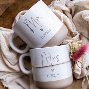 Op de afbeelding: Twee witte keramische mokken, &eacute;&eacute;n met de tekst "Mr." en de andere met "Mrs.", met een hartje en de datum "13.09.2025". De mokken hebben een gespikkeld ontwerp en een beige basis. E&eacute;n mok is versierd met een klein bloemstuk.