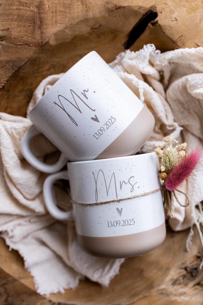 Op de afbeelding: Twee witte keramische mokken, &eacute;&eacute;n met de tekst "Mr." en de andere met "Mrs.", met een hartje en de datum "13.09.2025". De mokken hebben een gespikkeld ontwerp en een beige basis. E&eacute;n mok is versierd met een klein bloemstuk.