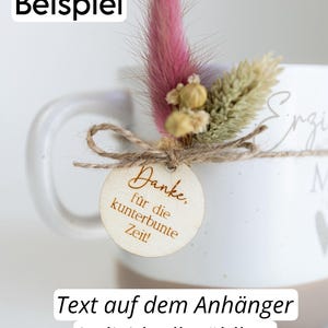 Op de afbeelding: Witte keramische mok met een arrangement van gedroogde bloemen en een houten label. Het label leest "Danke, f&uuml;r die kunterbunte Zeit!" en is vastgebonden met touw. De tekst "Text auf dem Anh&auml;nger individuell w&auml;hlbar" is ook zichtbaar.