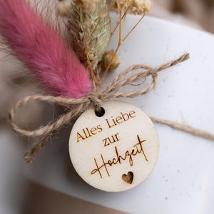 Op de afbeelding: Close-up van een geschenkdoos met een houten label en droogbloemen. Het label leest "Alles Liebe zur Hochzeit" met een hart. De bloemen omvatten een roze veerkruid en beige gedroogde sprietjes, vastgebonden met touw.