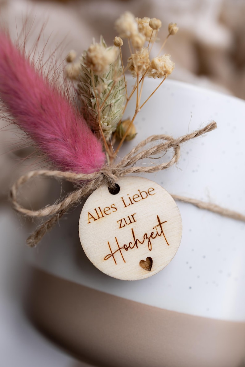 Op de afbeelding: Close-up van een geschenkdoos met een houten label en droogbloemen. Het label leest "Alles Liebe zur Hochzeit" met een hart. De bloemen omvatten een roze veerkruid en beige gedroogde sprietjes, vastgebonden met touw.