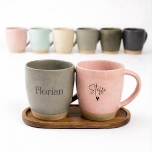 Könnte beinhalten: Eine Sammlung von Keramikbechern in verschiedenen Pastellfarben, darunter Rosa, Grün und Grau, mit einem naturfarbenen Boden. Zwei Becher im Vordergrund sind mit den Namen "Florian" und "Steffi" sowie einem Herzsymbol personalisiert. Die Becher stehen auf einem Holztablett.