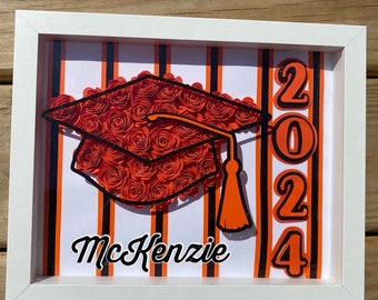 May include: A framed graduation shadow box featuring a mortarboard made of orange paper roses. The design includes the year 2024 in orange, black and white stripes, and the name "McKenzie" in cursive. The frame is white.