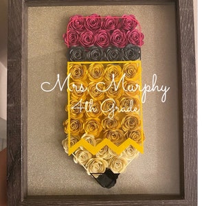 May include: A framed artwork featuring a pencil design made of paper roses. The top is pink, the middle is black, and the body is yellow and cream-colored. The pencil has the text "Mrs. Murphy 4th Grade" in white script.