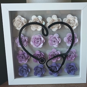 May include: A framed artwork featuring paper flowers in shades of cream, lavender, and purple. A black stethoscope shape is centered over the flowers, creating a heart shape. The frame is white.
