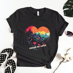 Op de afbeelding: Zwart T-shirt met een kleurrijke afbeelding van een hartvormig landschap met bergen, bomen en een skyline van een stad. De tekst "COLOR OF LOVE" is onder de afbeelding gedrukt.