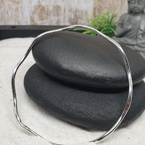May include: A silver-colored, wavy bangle bracelet. The bracelet is displayed on two black, smooth stones, set against a backdrop of white sand. A small Buddha statue and greenery are visible in the background.