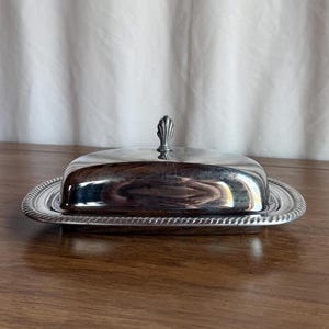 May include: A silver-plated butter dish with a domed lid and a decorative handle. The rectangular base has a rope-like border. The dish is displayed on a wooden surface.