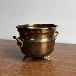 May include: A small, antique brass cauldron with three ornate feet and two handles. The cauldron has a rounded body and a wide rim. The brass has a warm, aged patina, giving it a vintage look. The cauldron sits on a wooden surface.