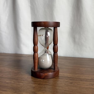 以下が含まれることがあります： 濃い茶色の木製フレームの砂時計。ガラス球には白い砂が入っています。砂時計は、4つの垂直な木製サポートで接続された丸いベースと上部によって支えられています。背景は白いカーテンです。
