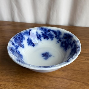 May include: A white and blue ceramic bowl with a floral pattern. The bowl has a scalloped edge and a detailed blue design around the rim and inside the bowl. The bowl is sitting on a wooden surface.