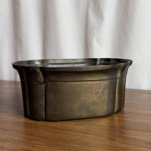 Può includere: Un contenitore rettangolare in ottone antico con bordo smerlato. I lati sono leggermente affusolati e il metallo ha un aspetto ossidato e invecchiato. Il contenitore è appoggiato su una superficie di legno.