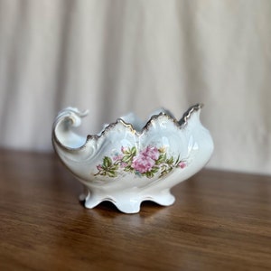 Puede incluir: Un tazón de porcelana blanca con un borde festoneado y un borde dorado. El tazón presenta un diseño floral con rosas rosadas y hojas verdes.