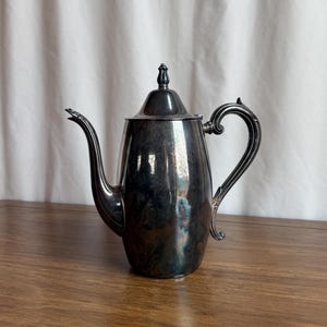 May include: A silver-colored coffee pot with a curved spout and handle. The pot has a lid with a decorative finial. The pot is sitting on a wooden surface.