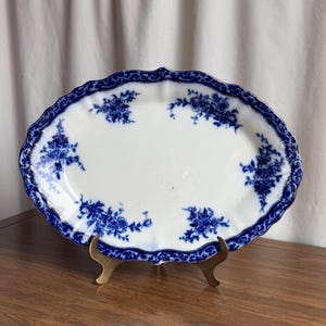 May include: An oval, antique serving platter with a white center and a blue floral design. The rim is scalloped and features a detailed blue pattern. The platter is displayed on a gold-colored stand.